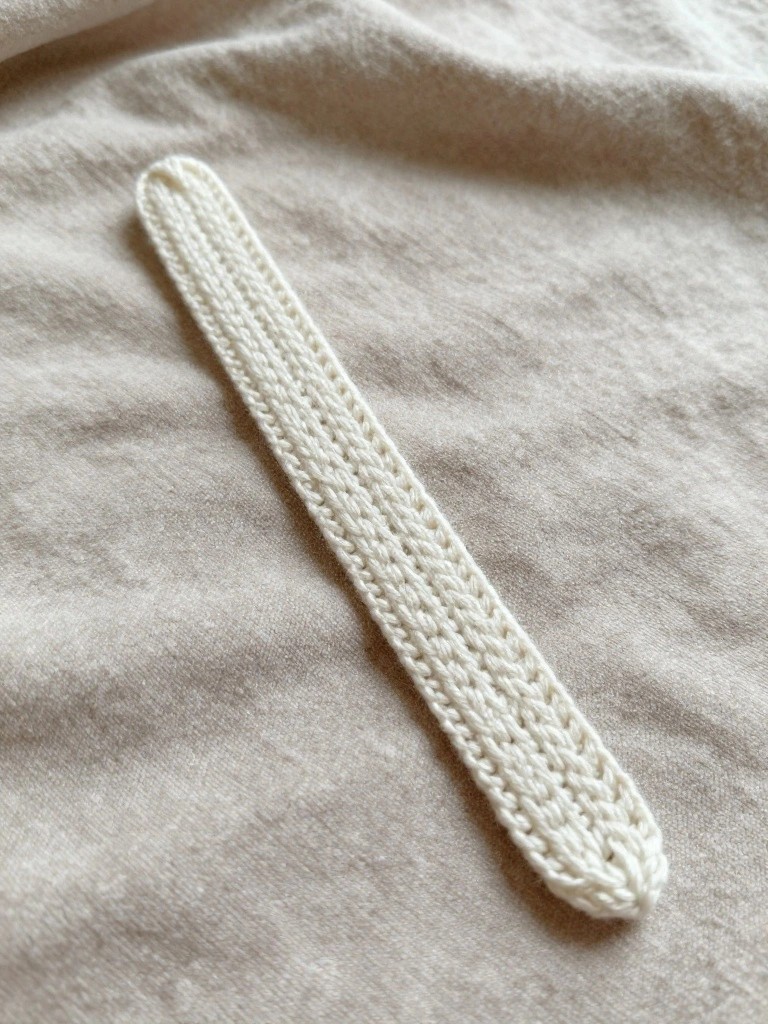 A long narrow white knitted bookmark with braided texture lies flat on beige fabric.