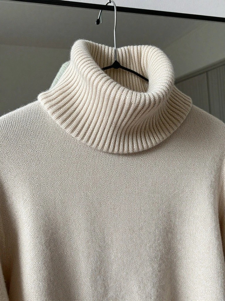 Cream-colored knitted turtleneck sweater with ribbed collar hanging on a black hanger against a mirror.