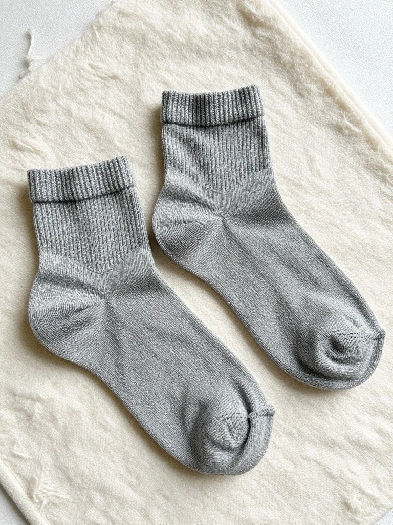 A pair of light gray knitted ankle socks with ribbed cuffs and side texture laid flat on beige fabric.