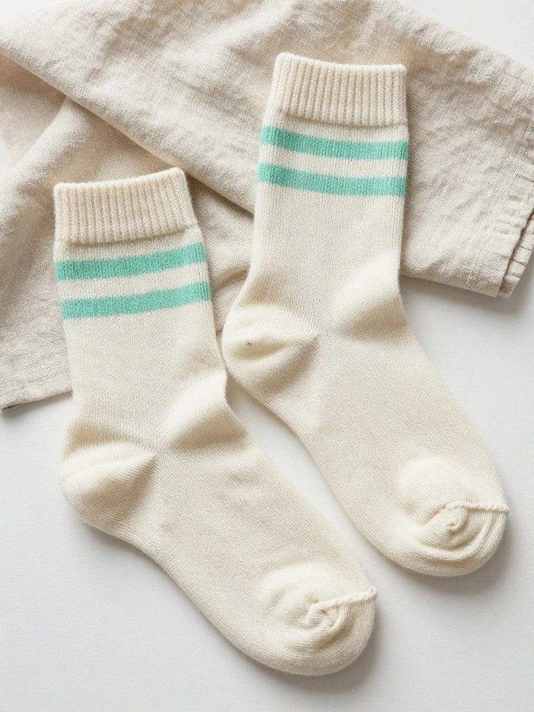 Two cream knitted crew socks with mint green stripes below ribbed cuffs lie flat on a white surface draped with beige linen cloth.