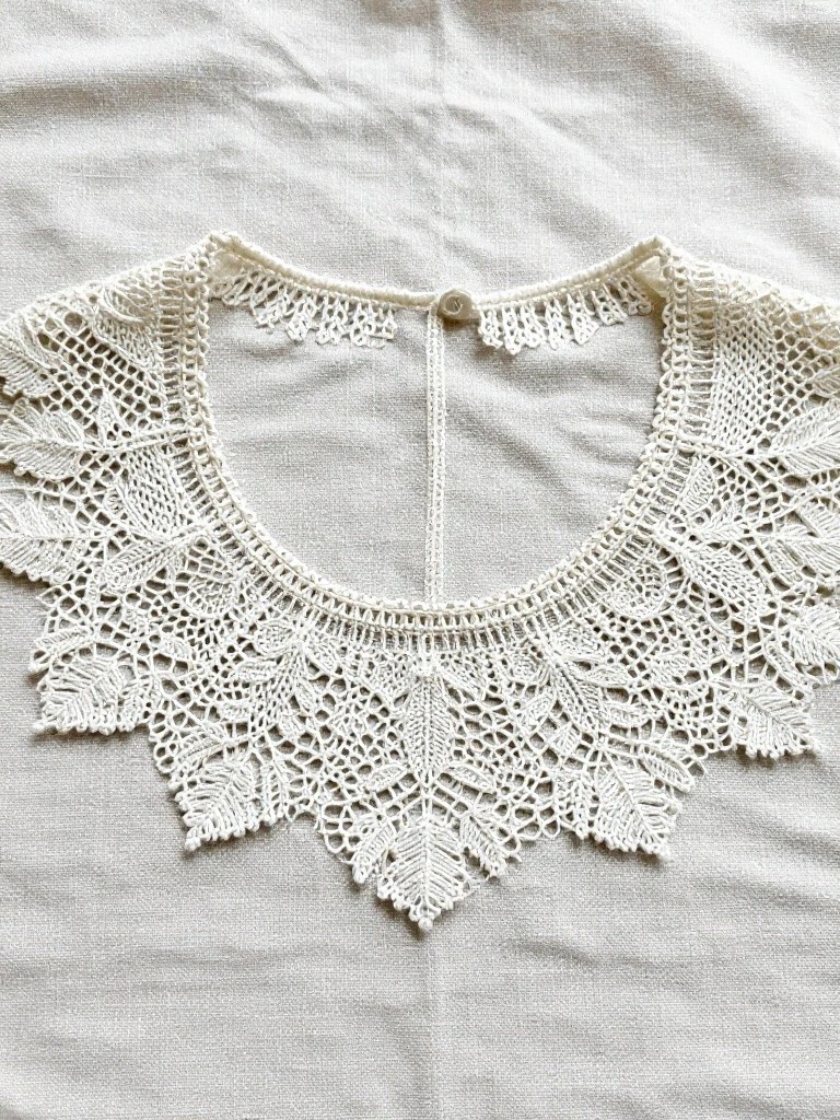 White knitted lace collar with leaf-shaped motifs and scalloped edges, laid flat on fabric with a button at the open back.