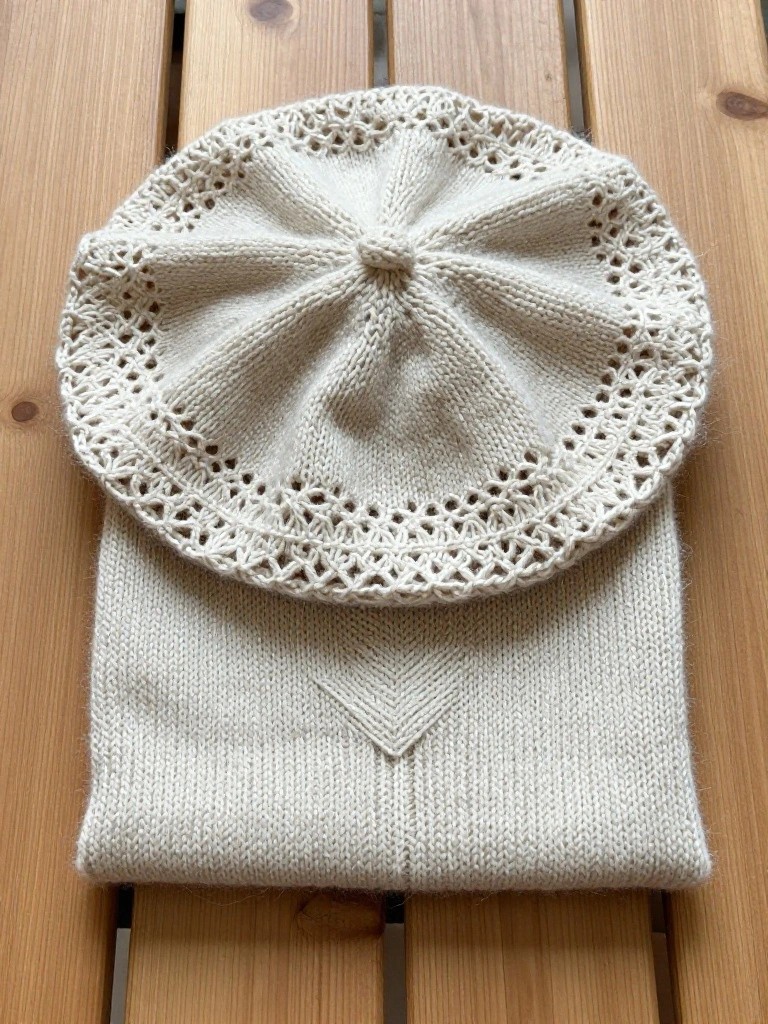 Cream knitted beret hat with wide lacy brim on top of matching rectangular cowl with front V detail on wood surface.
