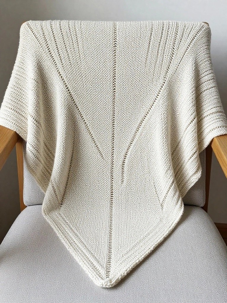 A cream-colored knitted triangular shawl with open lace patterns draped over the back of a wooden chair.