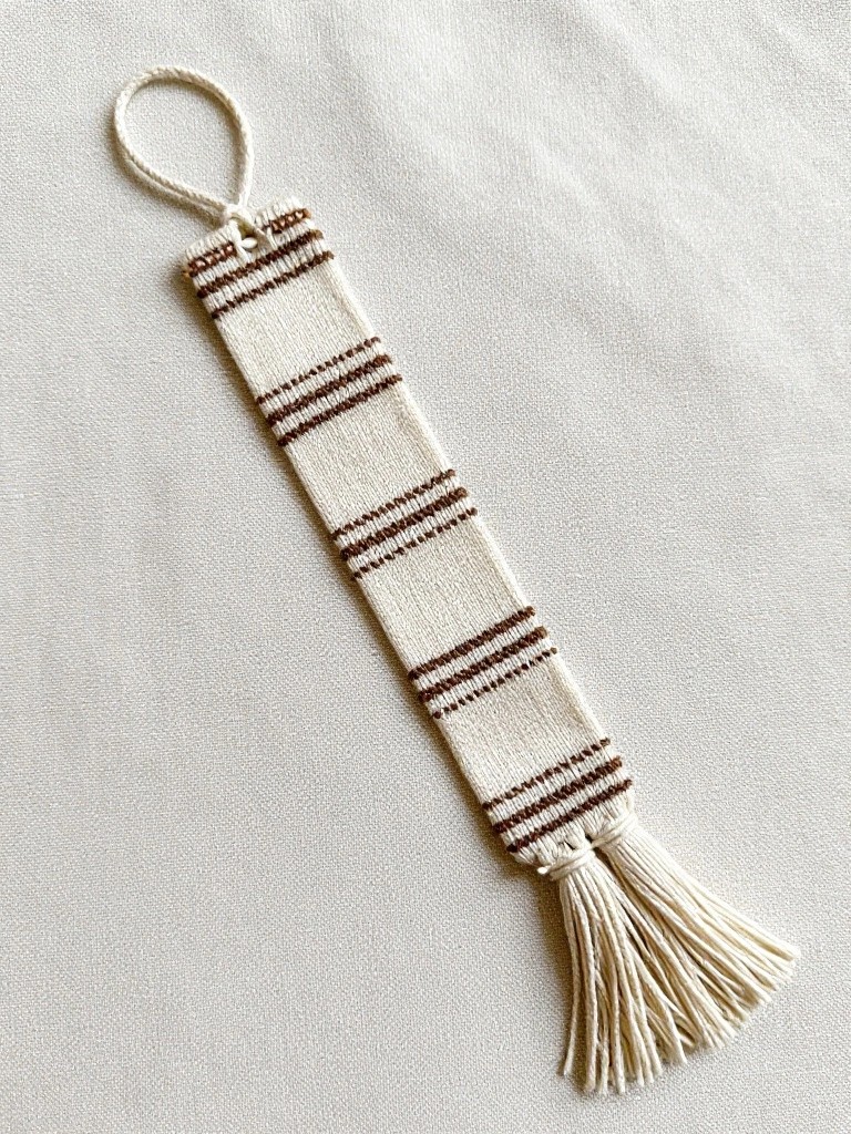 Knitted bookmark with cream and brown horizontal stripes, top loop, and bottom white tassels on white background.