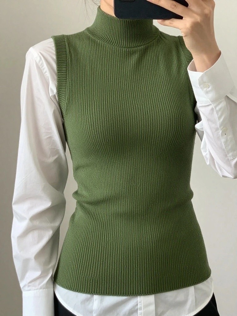 A woman takes a mirror selfie wearing an olive green ribbed knit sleeveless turtleneck vest over a white long-sleeve button-up shirt.