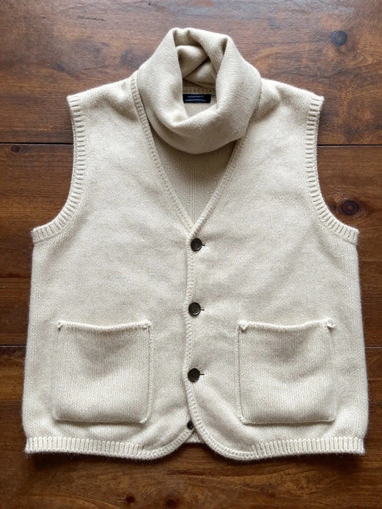 Cream-colored sleeveless knitted vest with turtleneck collar, three front buttons, and two patch pockets on a wooden table.