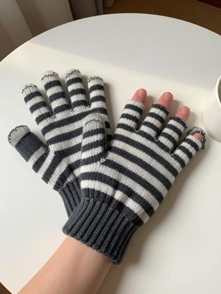 Two pairs of black and white horizontally striped fingerless knitted mitts with ribbed cuffs lie flat on a white table next to a cup.