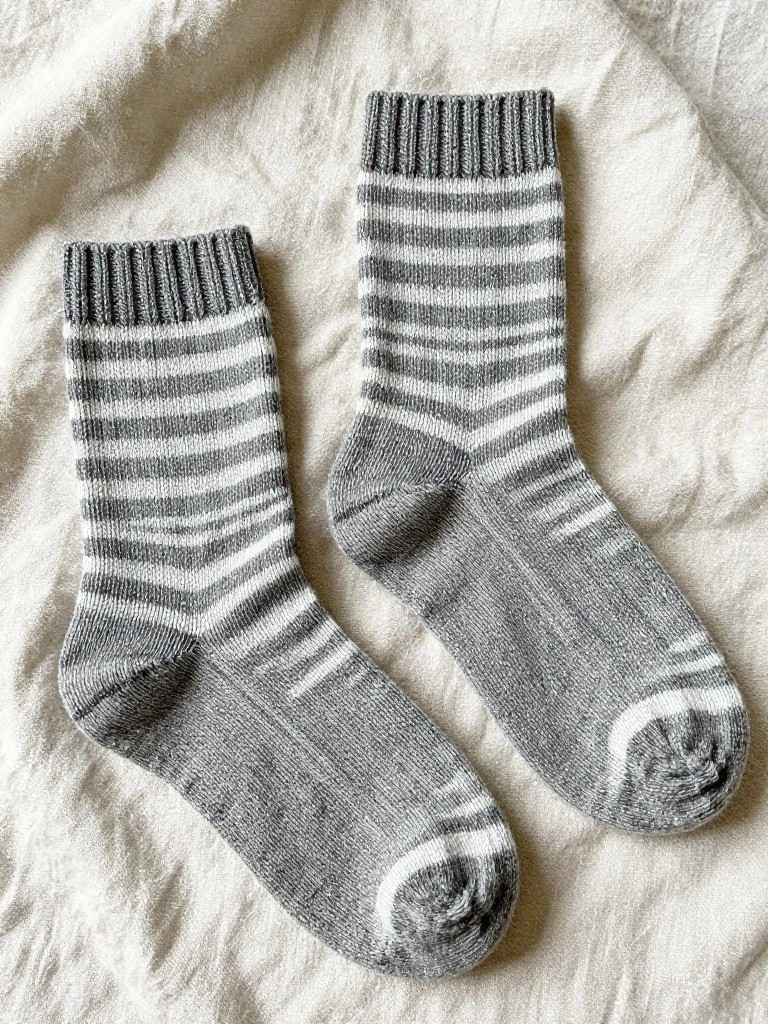 A pair of gray knitted socks with white zigzag stripes and ribbed cuffs, laid flat on beige fabric.