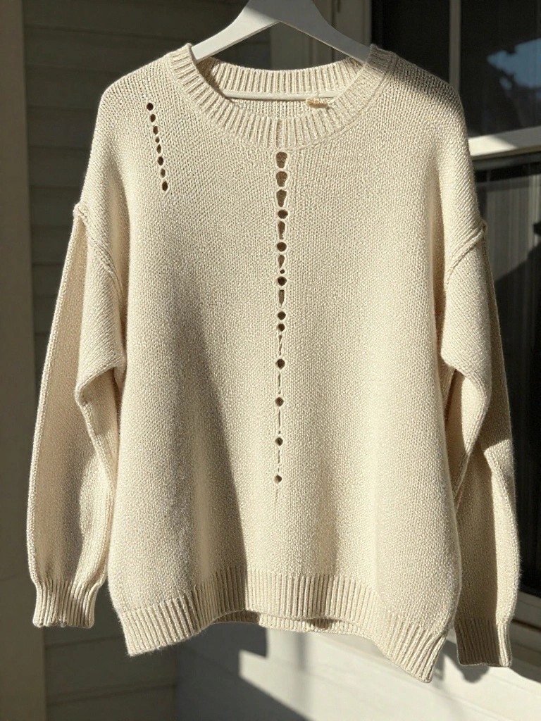 Cream-colored oversized knitted sweater with round holes down the center front and on the shoulders, hanging on a white hanger by a window.