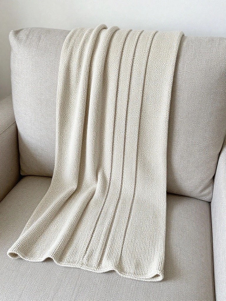 Cream-colored knitted throw blanket with vertical ribs draped over a beige couch arm.