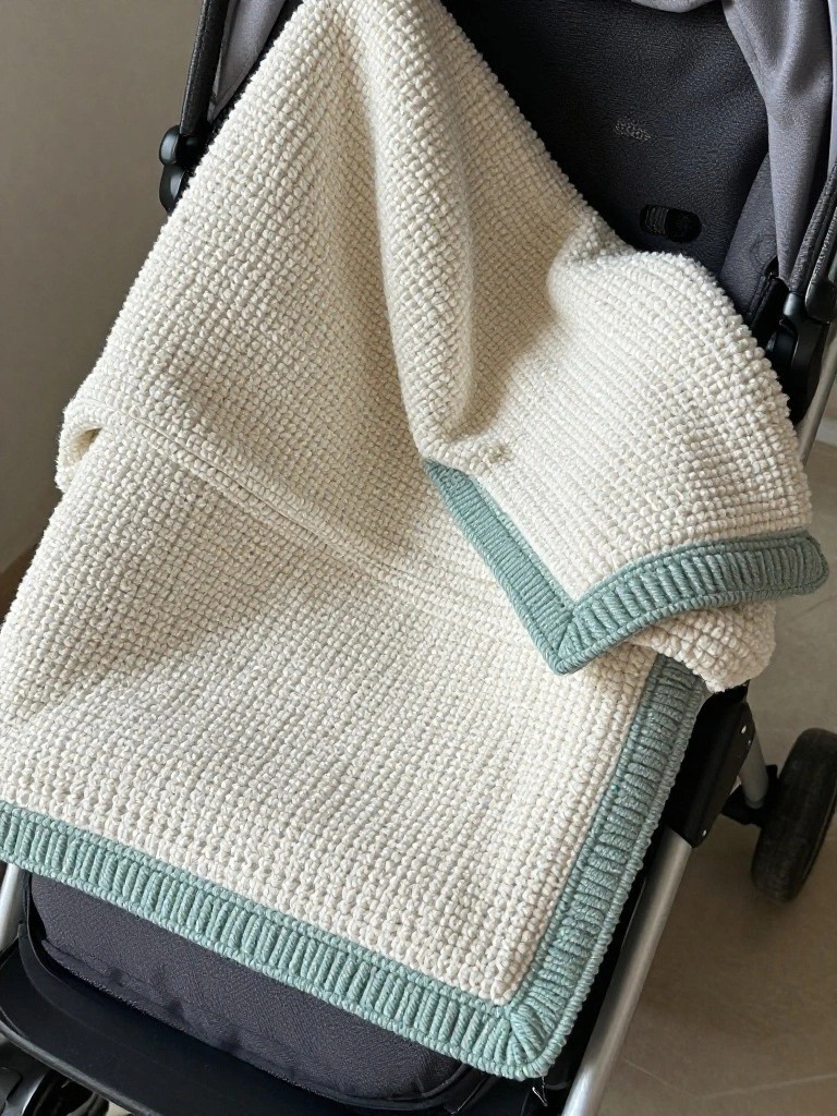 A cream knitted blanket with teal edging draped over a black stroller.