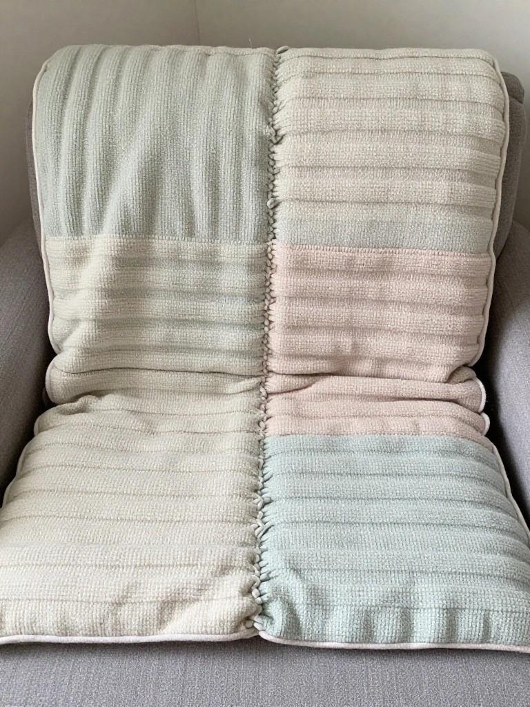 A square knitted blanket made of four fringed panels in gray, beige, pink, and mint with horizontal textured stripes, draped over a gray chair.