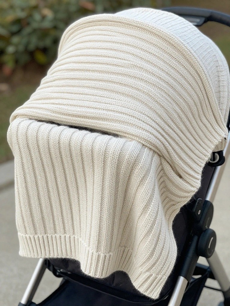 A cream-colored chunky ribbed knit blanket covers the hood and seat of a black stroller.