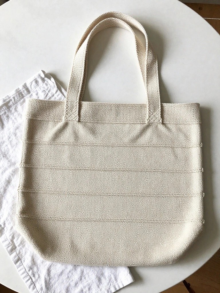 A beige knitted tote bag with horizontal ridges and thick strap handles sits on a white table next to a folded white napkin.
