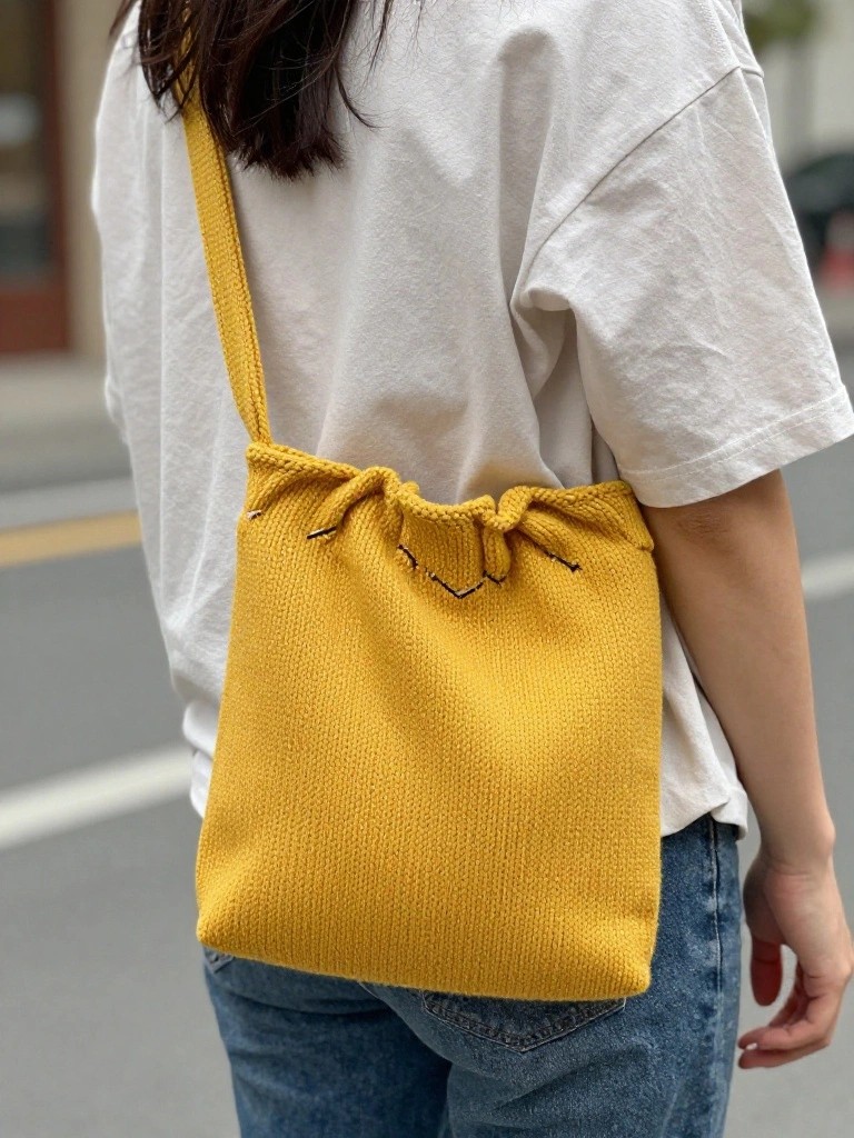 Back view of a woman in white t-shirt and jeans wearing a mustard-yellow knitted shoulder bag with black drawcord ties at the gathered top.