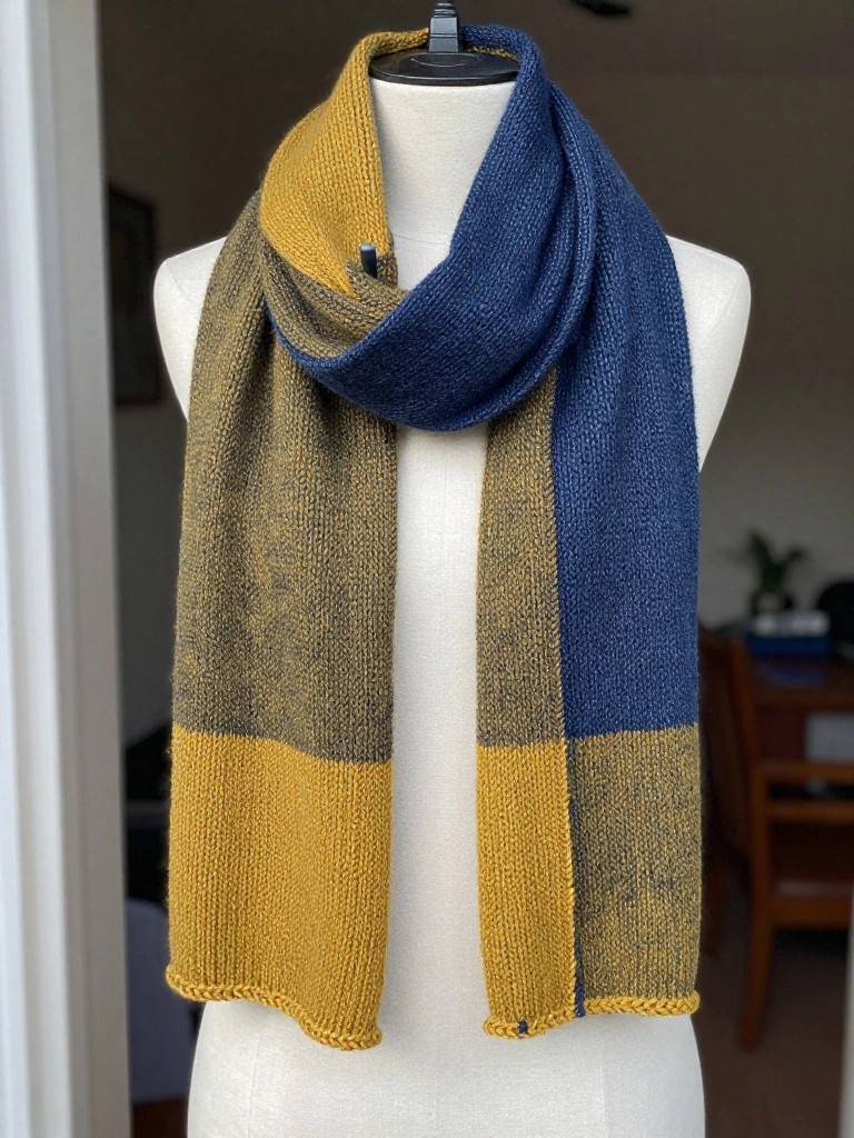A long knitted scarf with color-blocked panels in mustard yellow, olive green, and navy blue draped around a white mannequin's neck.