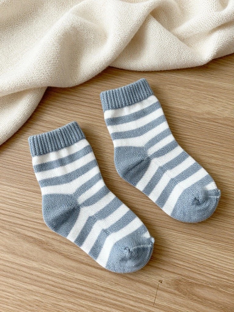 Two small light blue and white striped knitted baby ankle socks with ribbed cuffs on a wood surface near white fabric.
