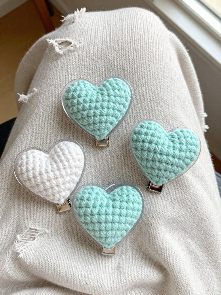 Four small crocheted hearts in white and mint green attached to metal clips on a white knit fabric.