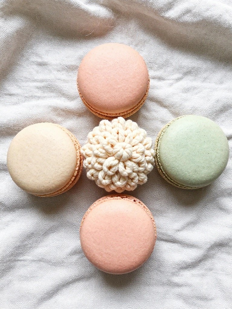A cream-colored crocheted looped flower sits among four pastel macarons on white fabric.