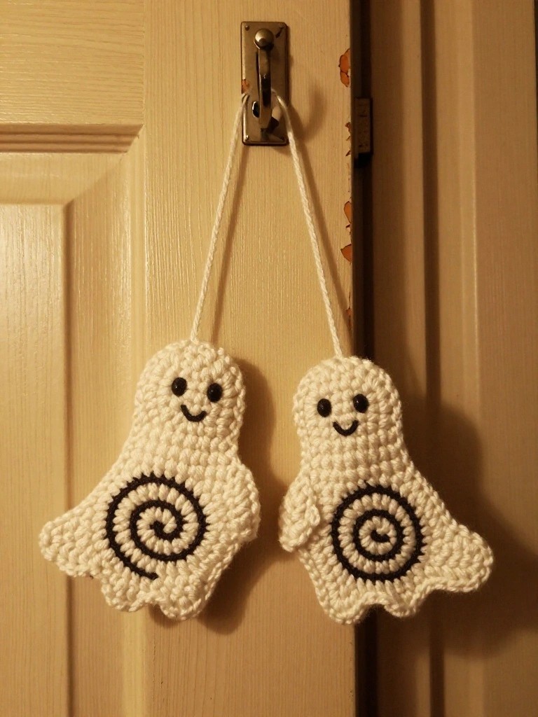 Two white crocheted ghost shapes with black spirals hang from a door handle by white yarn strings.