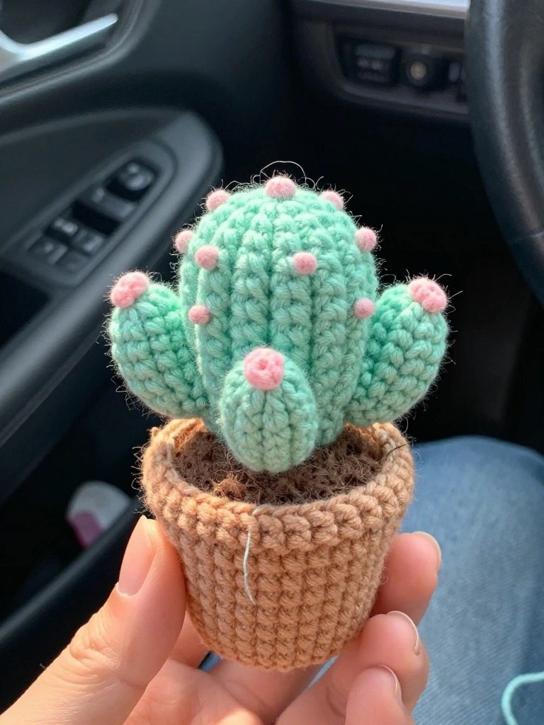 Hand holding a mint-green crocheted cactus with pink accents in a brown pot.
