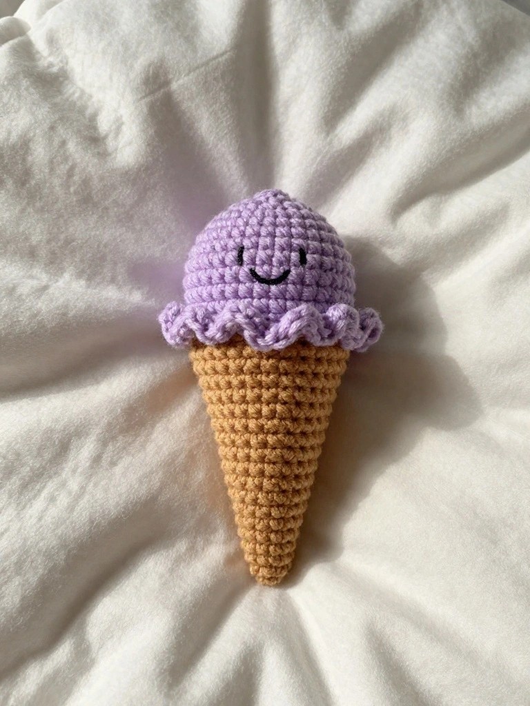 Crocheted purple ice cream cone with smiling face on white fabric.