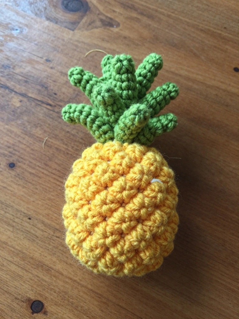 Crocheted yellow pineapple with green leafy top on wooden surface.
