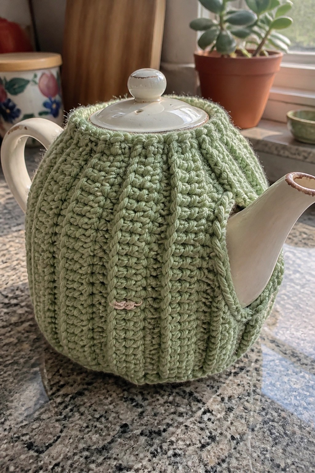 Light green cable-knit tea cozy on white ceramic teapot atop granite counter