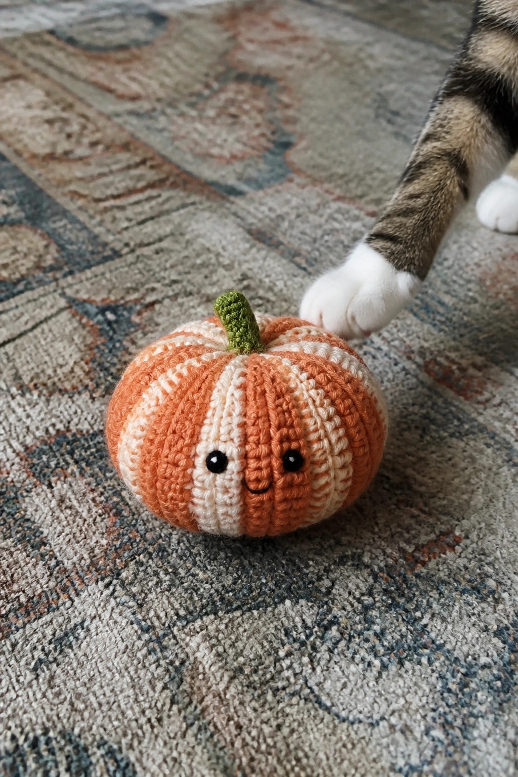 Crocheted orange-white striped pumpkin with smiling face, cat paw nearby on rug.