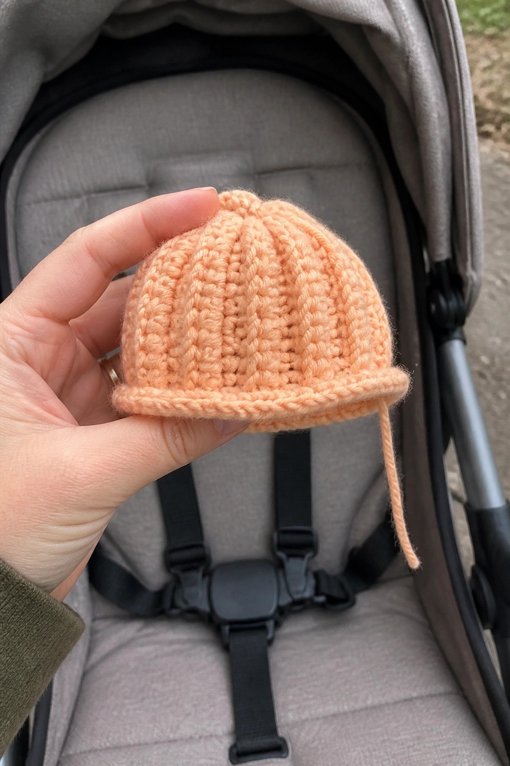 Hand holding small peach knitted baby hat over gray stroller with harness