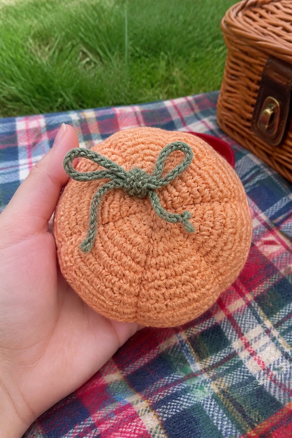 Hand holding crocheted orange pumpkin with green bow on plaid picnic blanket near basket
