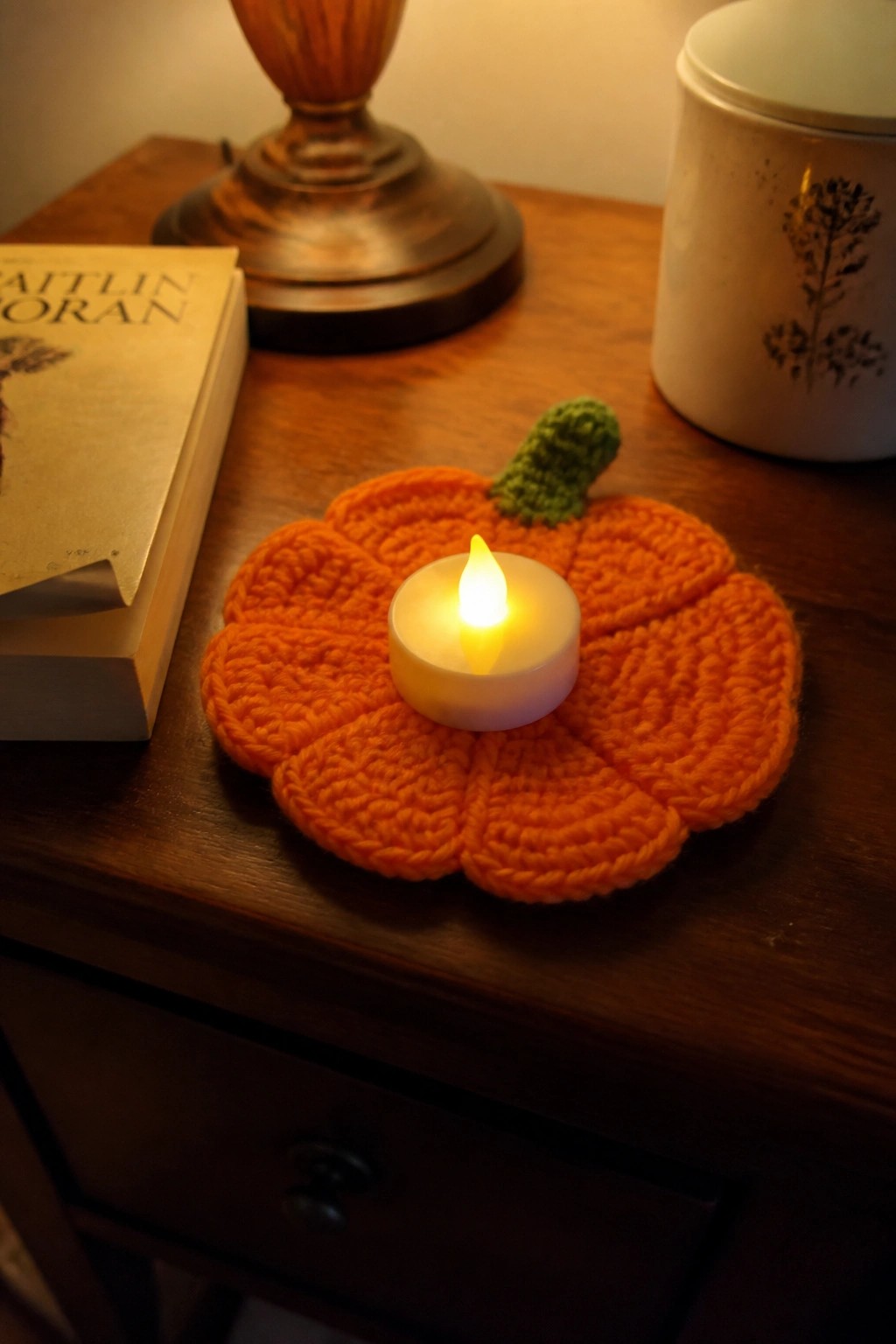 Lit tea light on orange crocheted pumpkin coaster atop wooden table beside book