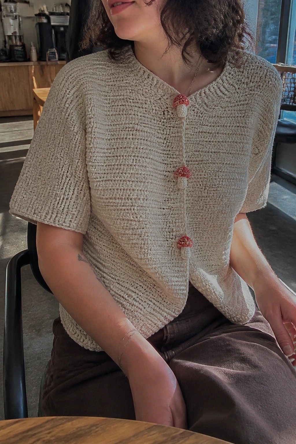 Beige crocheted cardigan with red mushroom buttons on a person in a cafe.