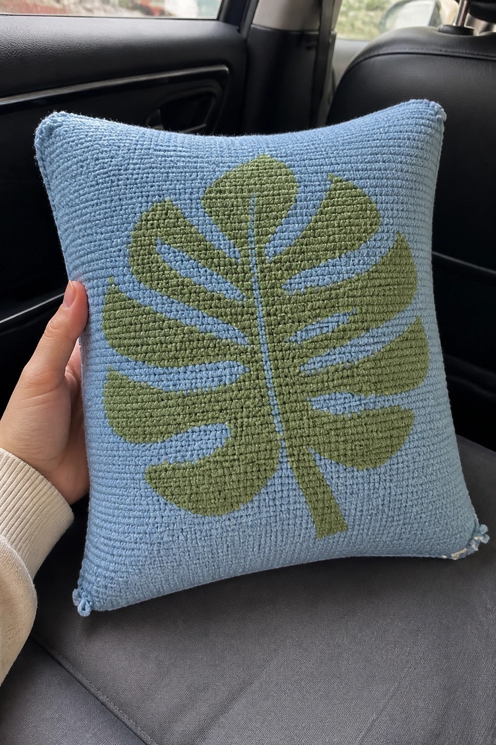 Hand holding light blue crocheted pillow with green leaf design on car seat.
