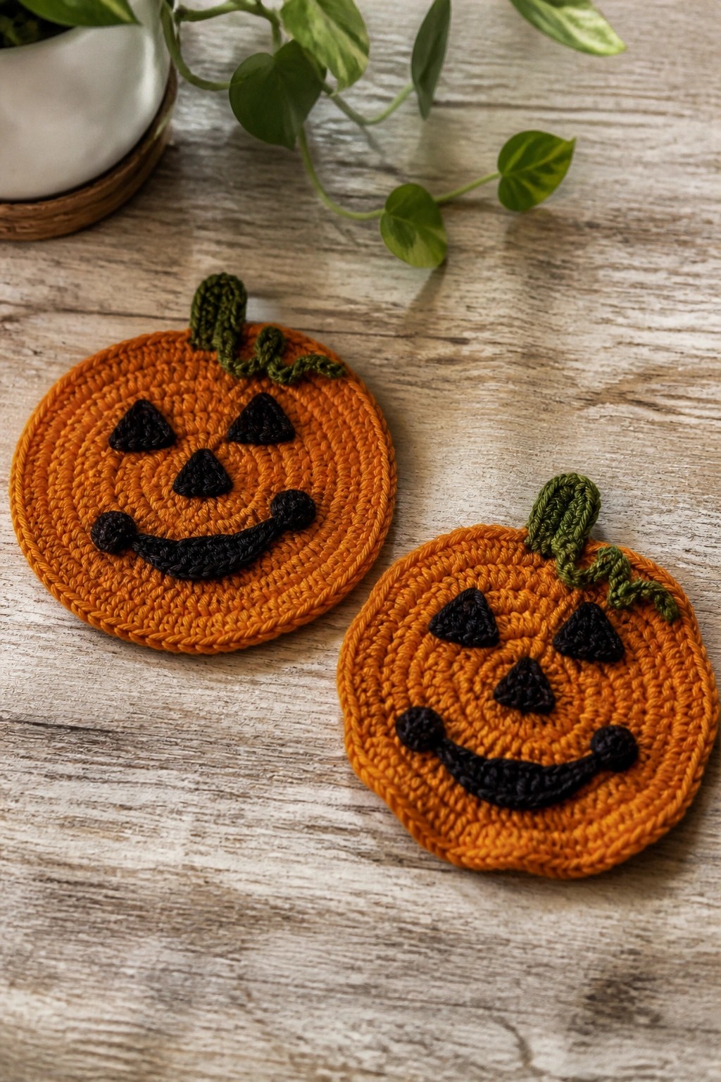 Two orange crocheted coasters with black jack-o'-lantern faces and green stems on a wooden table.