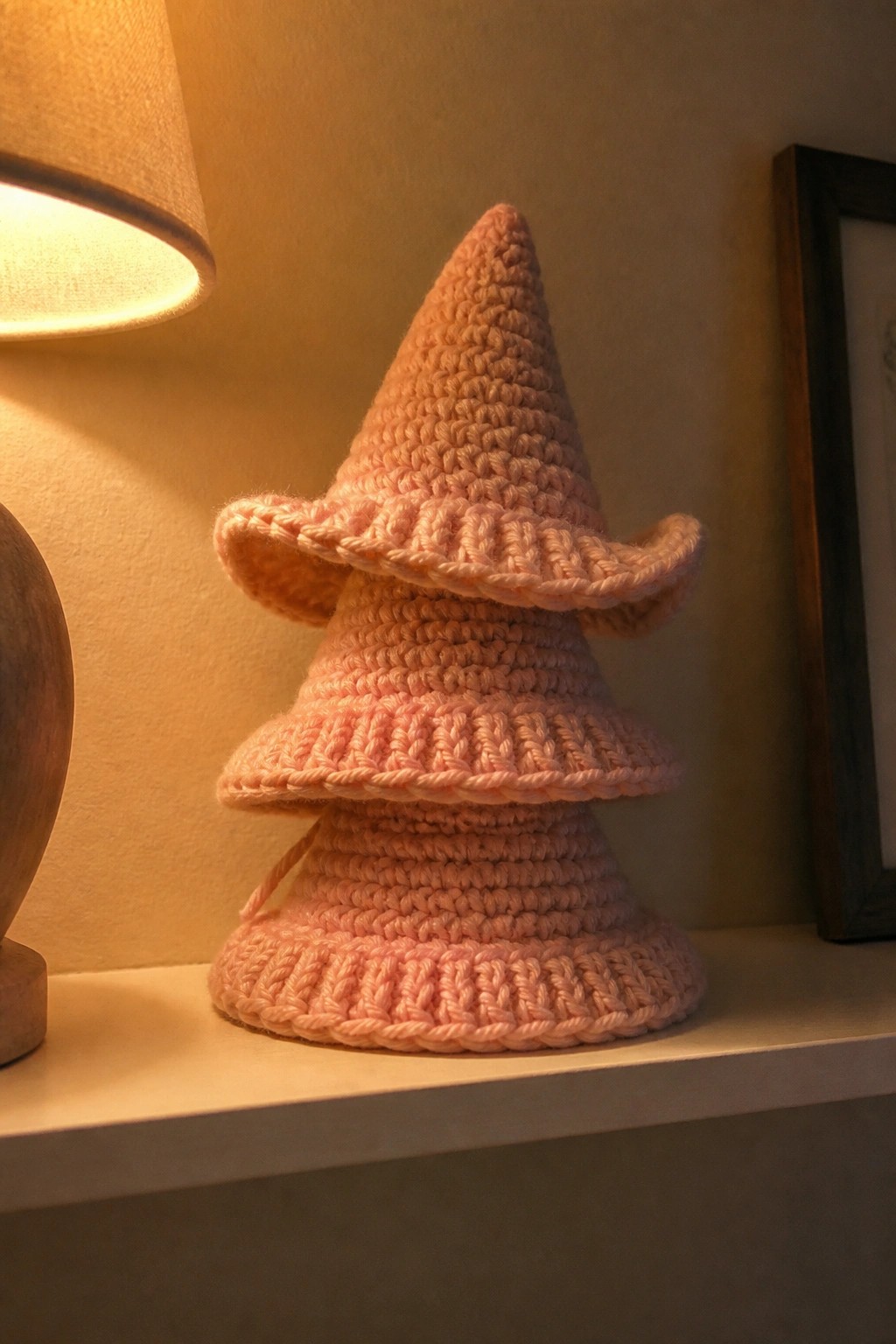 Pink crocheted tiered witch hat with ruffled brims on a shelf under warm light