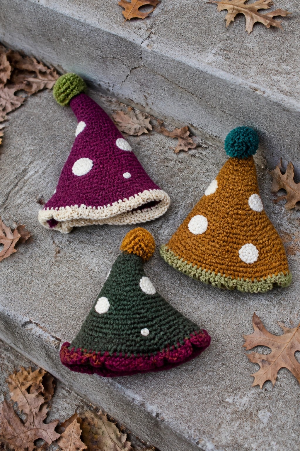 Three crocheted party hats with polka dots and pom-poms on concrete steps with leaves