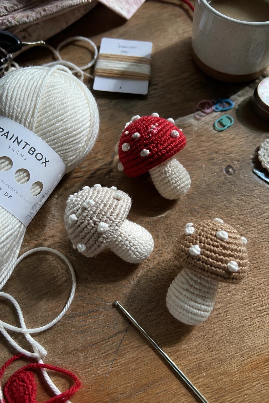 Three crocheted mushrooms in red and beige with yarn, hook, and supplies on wood.