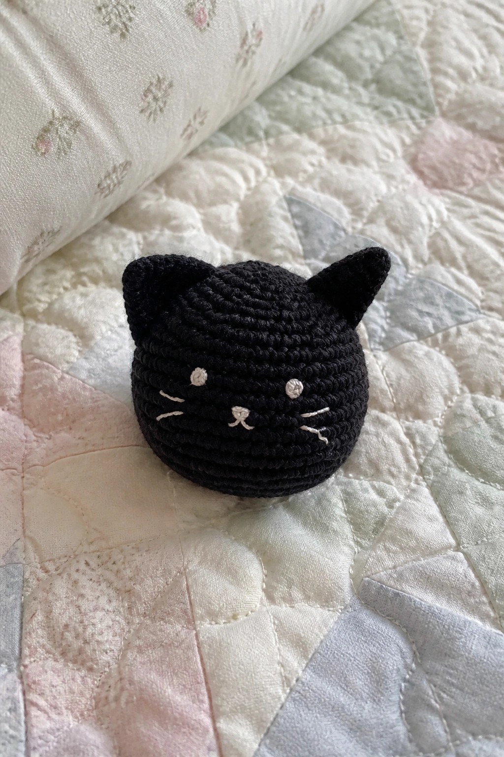 Black crocheted cat head with white embroidered face on pastel patchwork quilt