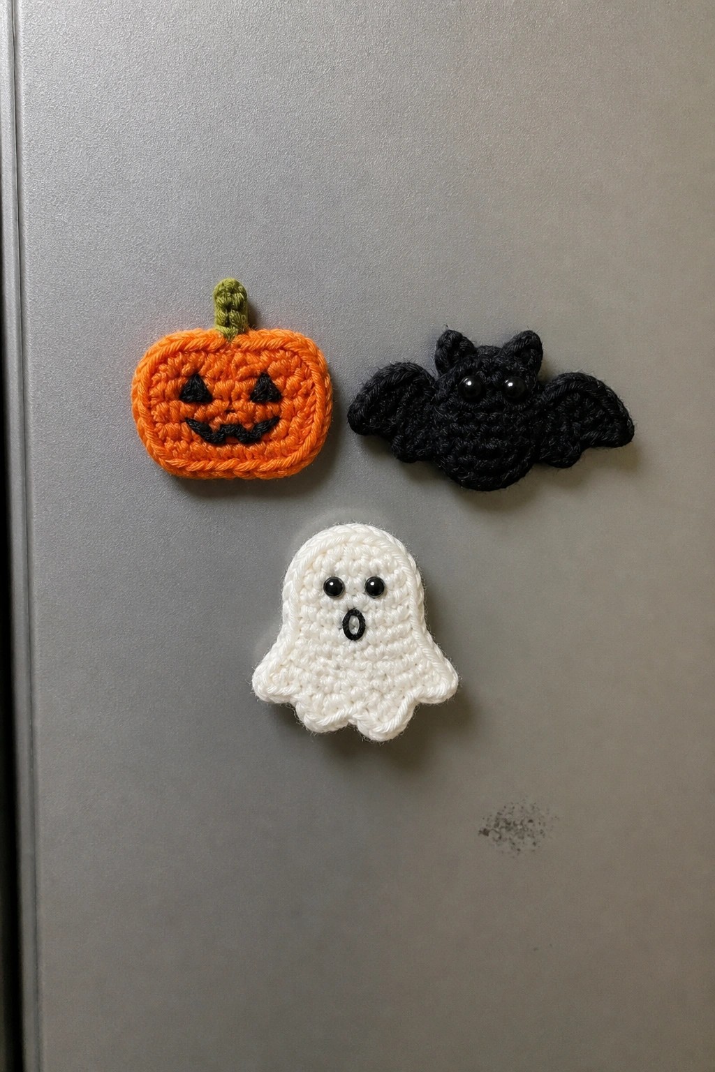 Crocheted orange pumpkin, black bat, and white ghost magnets on a refrigerator door.