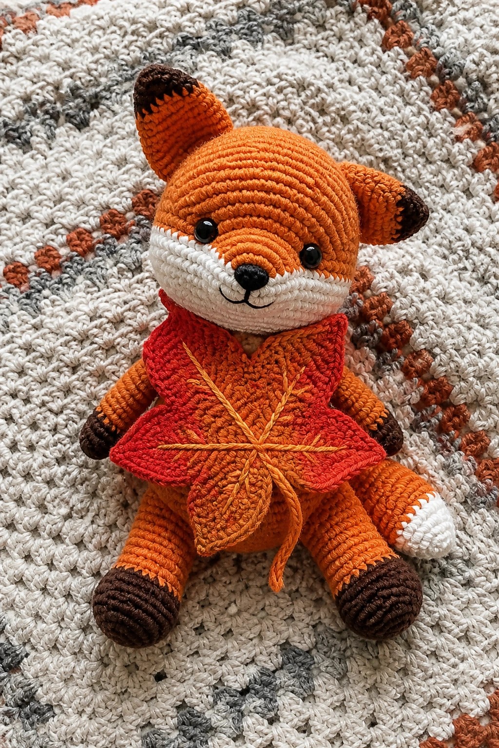 Orange crocheted fox wearing red leaf poncho on multicolored knitted blanket