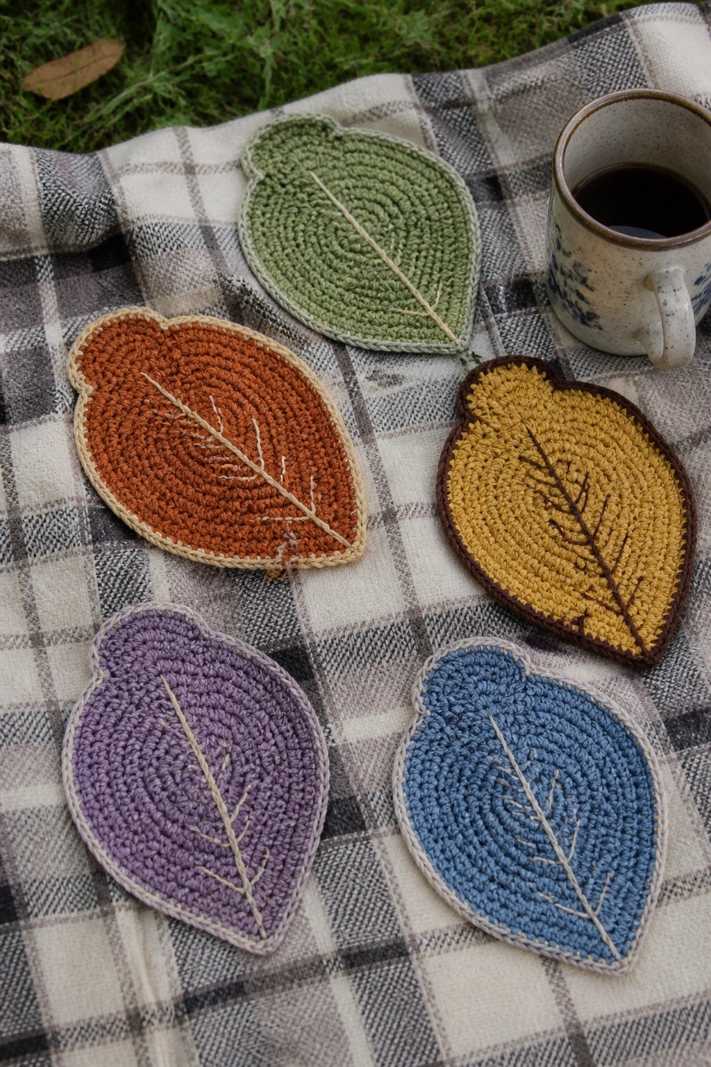 Colorful crocheted leaf coasters on plaid blanket beside coffee mug outdoors