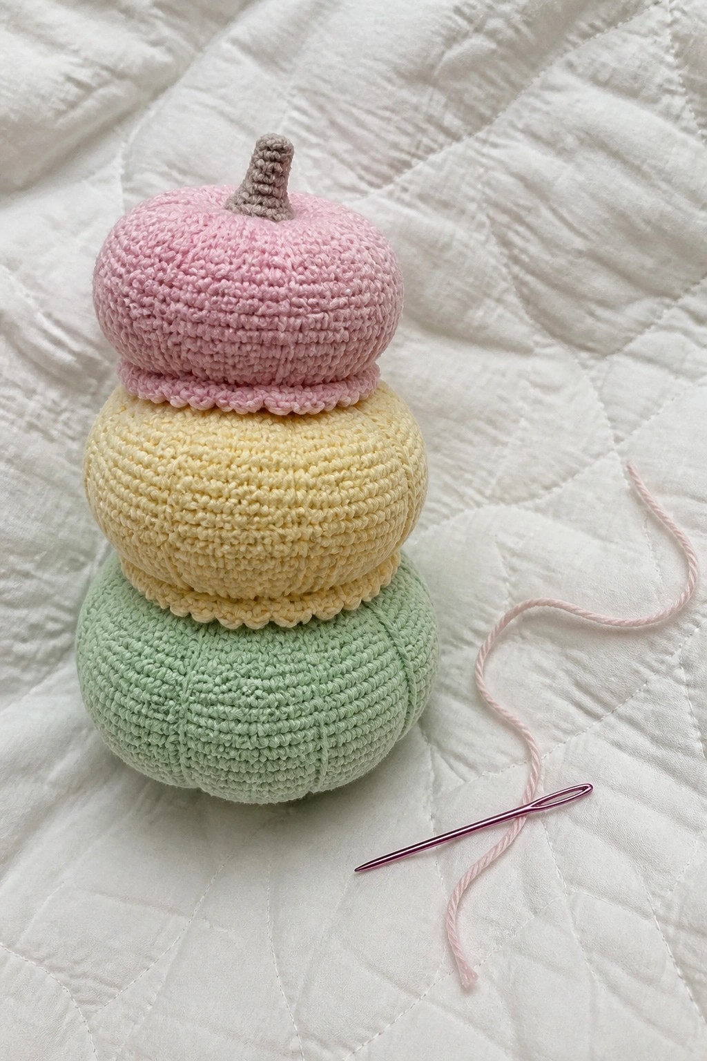 Stacked crocheted pastel pumpkins beside pink yarn and needle on white quilt
