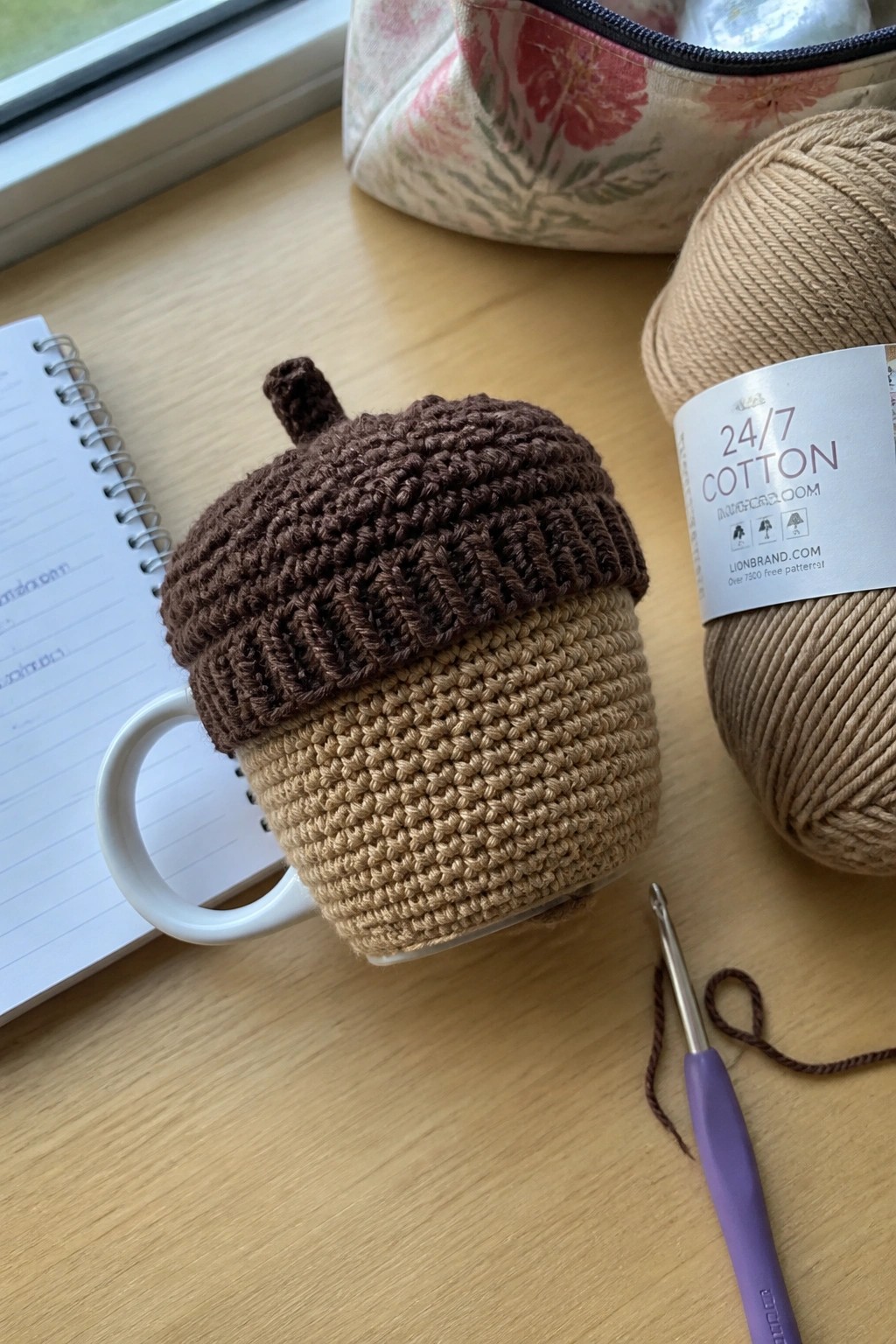 Crocheted acorn cozy on white mug beside beige yarn ball and purple hook