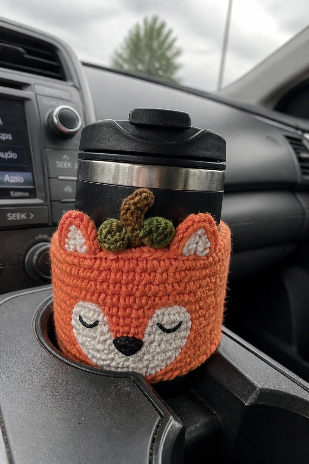 A crocheted orange fox cozy with a white face and small pumpkin topper covers a black travel mug in a car cup holder.