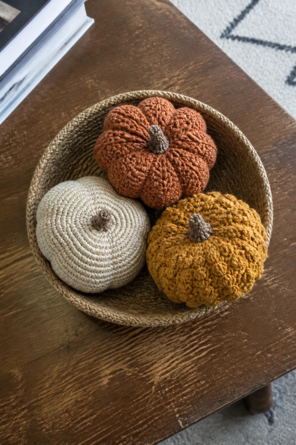 Three crocheted pumpkins in cream, rust, and mustard inside a woven basket on wood