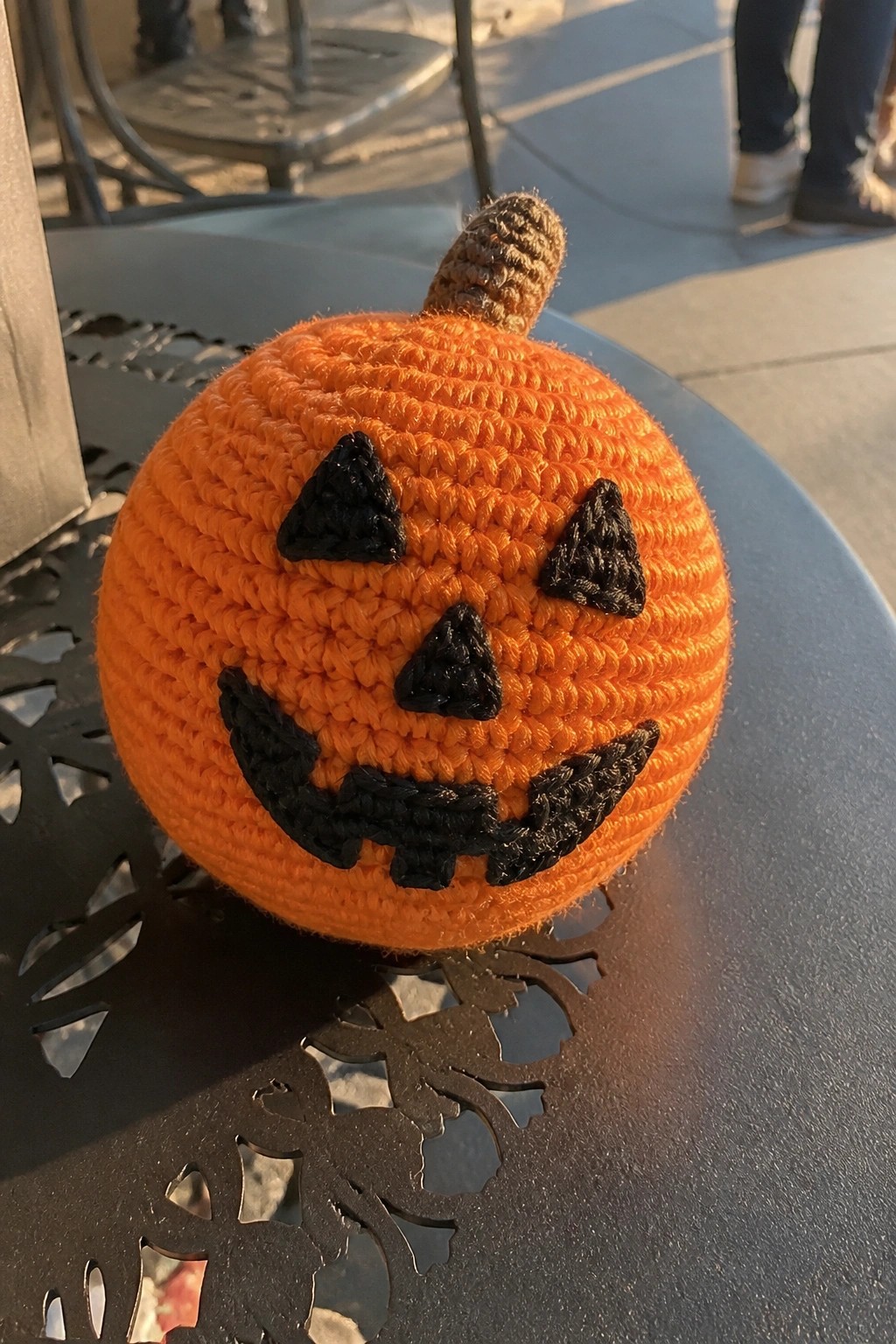 A small crocheted orange pumpkin with black triangular eyes and a jagged mouth sits on a dark metal table outdoors.