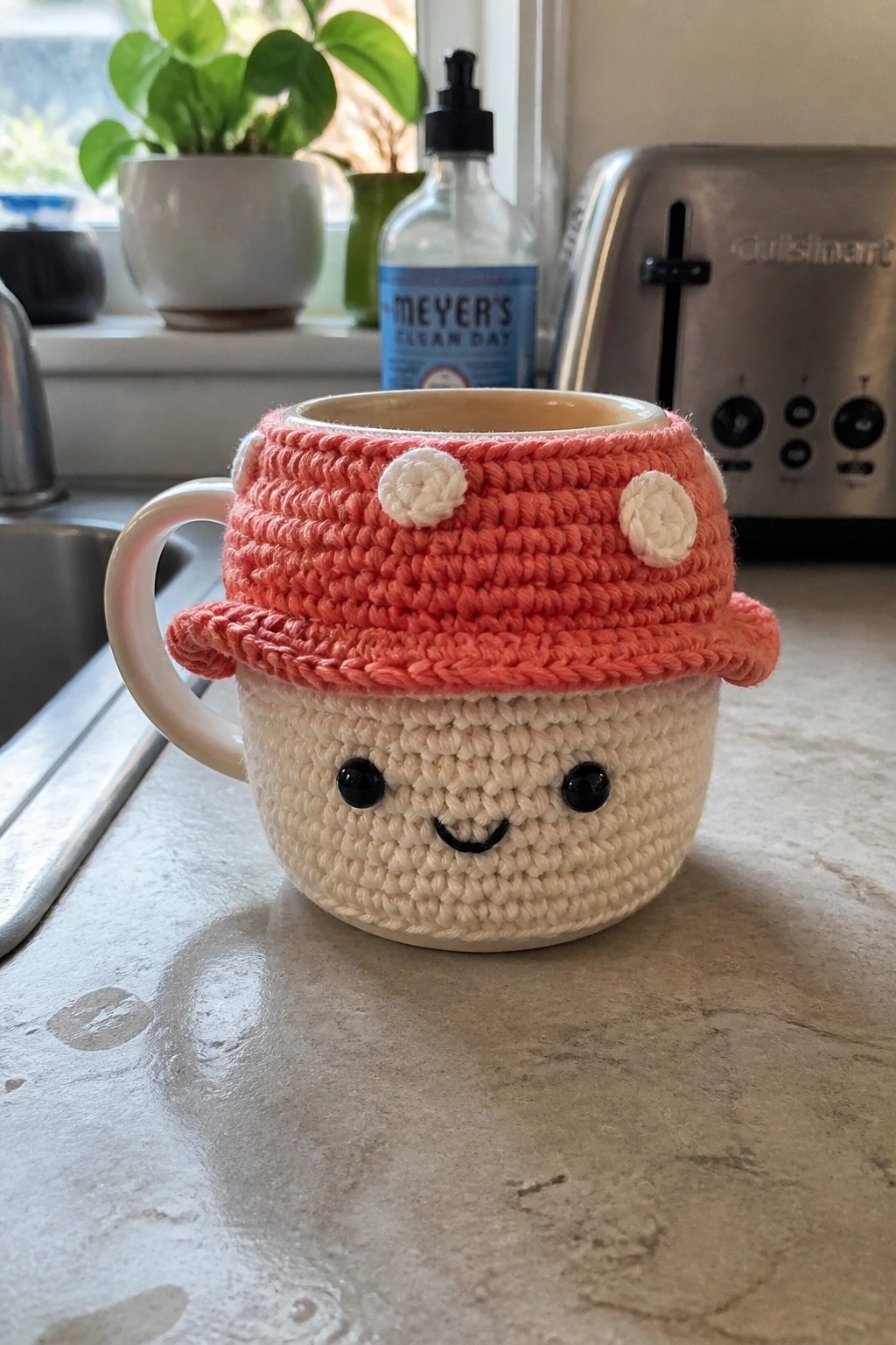 Pink crocheted mug cozy with smiling face and polka-dot hat on kitchen counter.
