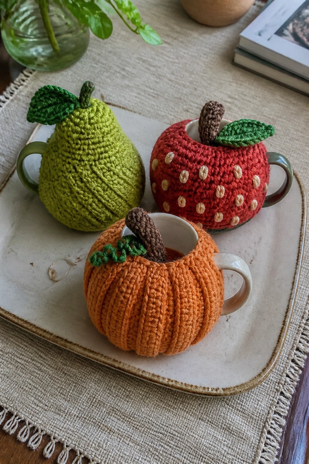 Crocheted green pear, red apple, and orange pumpkin cozies on mugs on tray