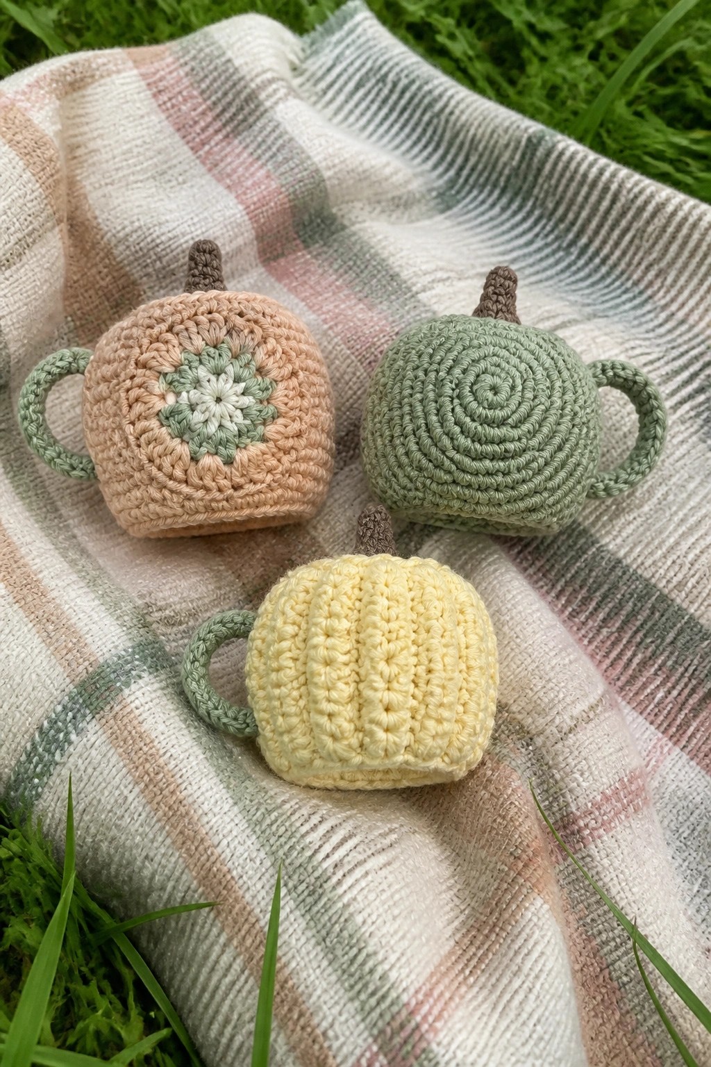 Three small crocheted pumpkin-shaped cozies with handles sit on a plaid blanket.